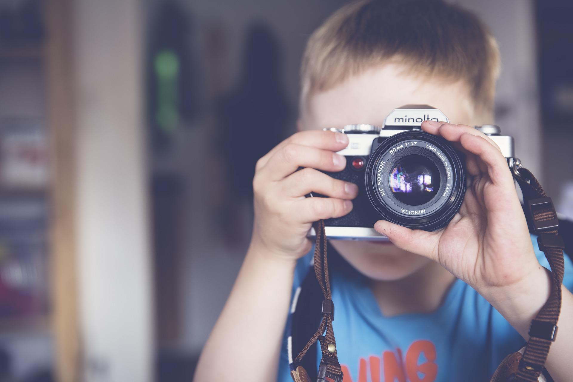 Junges Kind mit kurzen Haaren, hat einen Fotoapparat vor dem Gesicht und fotografiert den Zuschauer