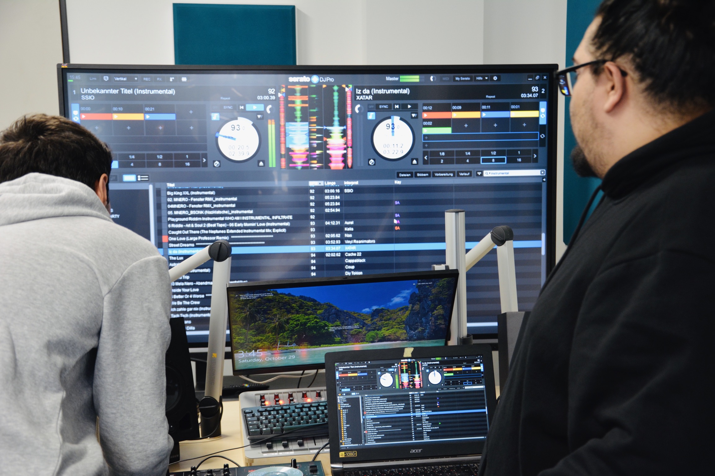 Zwei Männer in einem Radiostudio vor einem großen Bildschirm mit DJ Software
